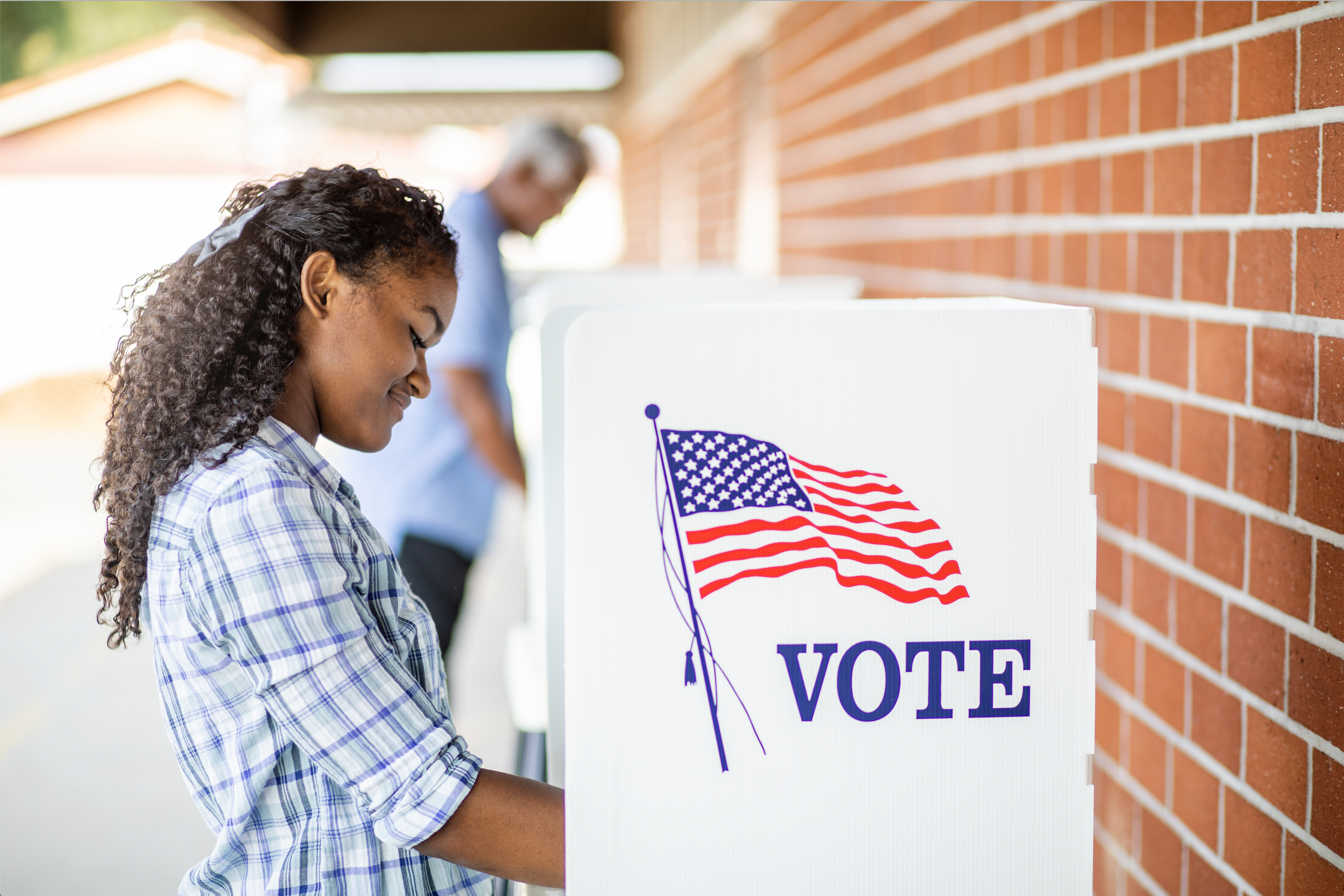 Young black woman voting at ballot box next to brick wall.